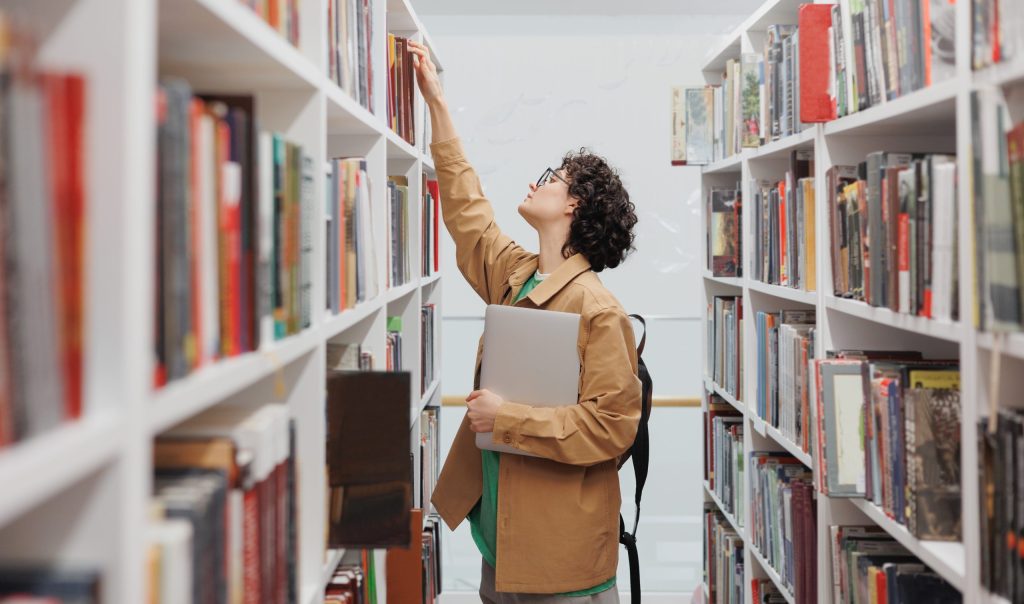young woman with curly hair in library among shelves with books. female student conducts scientific work
